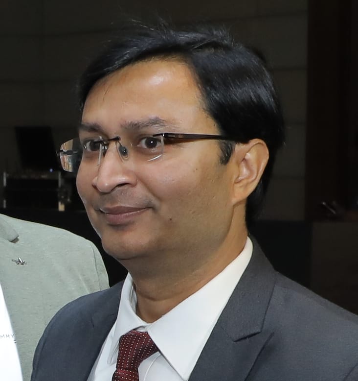 A man with short black hair and glasses, wearing a gray suit, white shirt, and red tie, stands indoors against a blurred global store background.